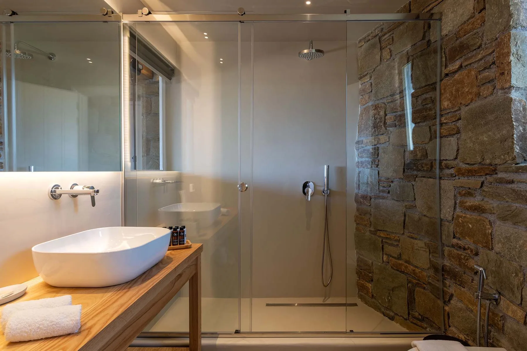 A bathroom with a glass shower, stone wall, and countertop basin.
