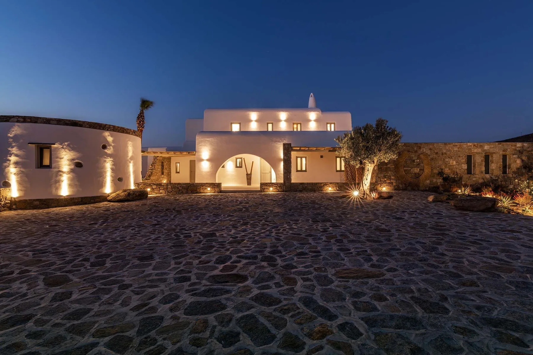 A warmly lit building stands beyond a broad stone-paved courtyard at dusk.