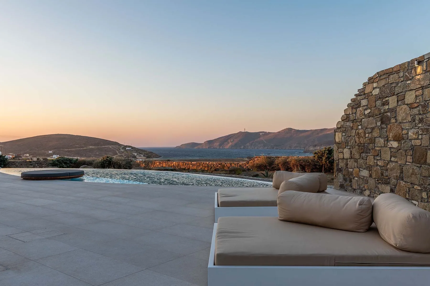A terrace with cushioned seating overlooks a pool and the sea at sunset.