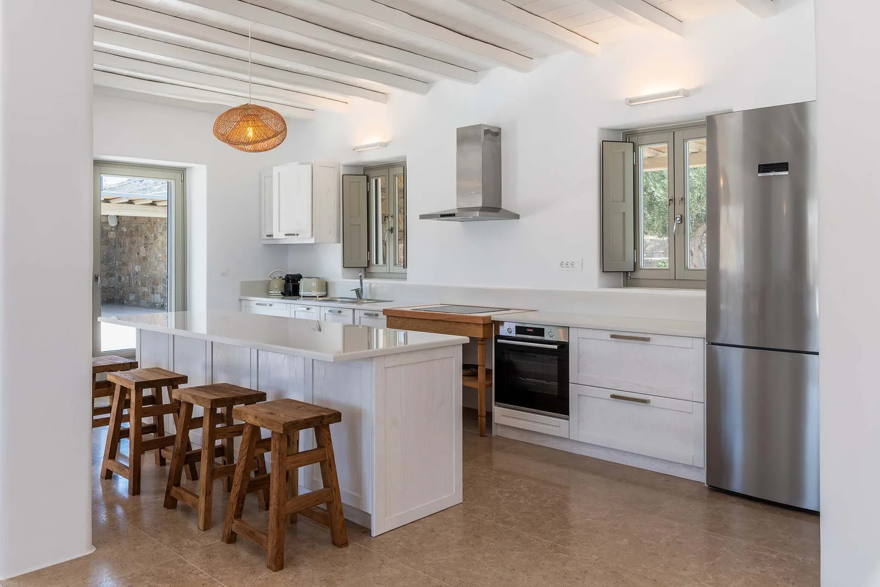 A bright kitchen with a central island, wooden stools, and stainless steel appliances.