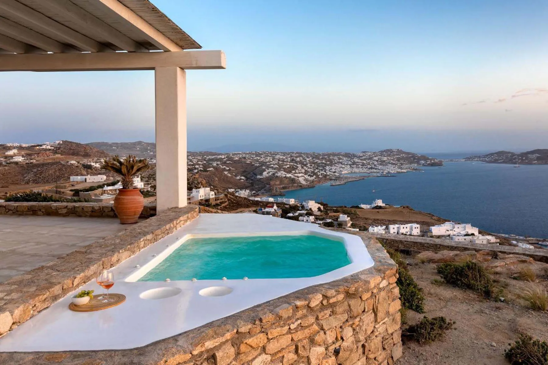 A hillside terrace overlooks the sea, with a turquoise pool beside a shaded seating area.