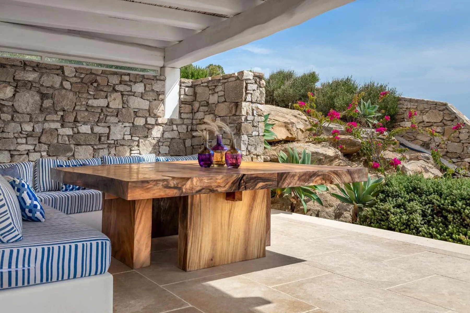 A covered outdoor dining area with a wooden table and striped seating overlooks a garden.
