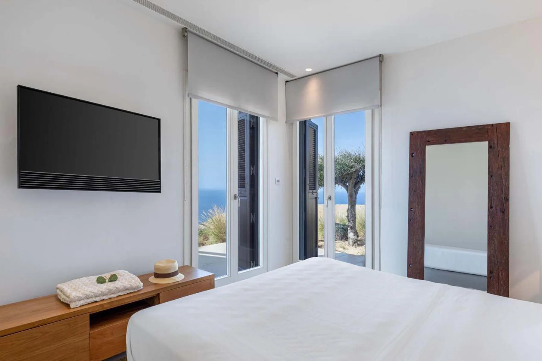 A bright bedroom with a bed, wall-mounted television, and large windows.