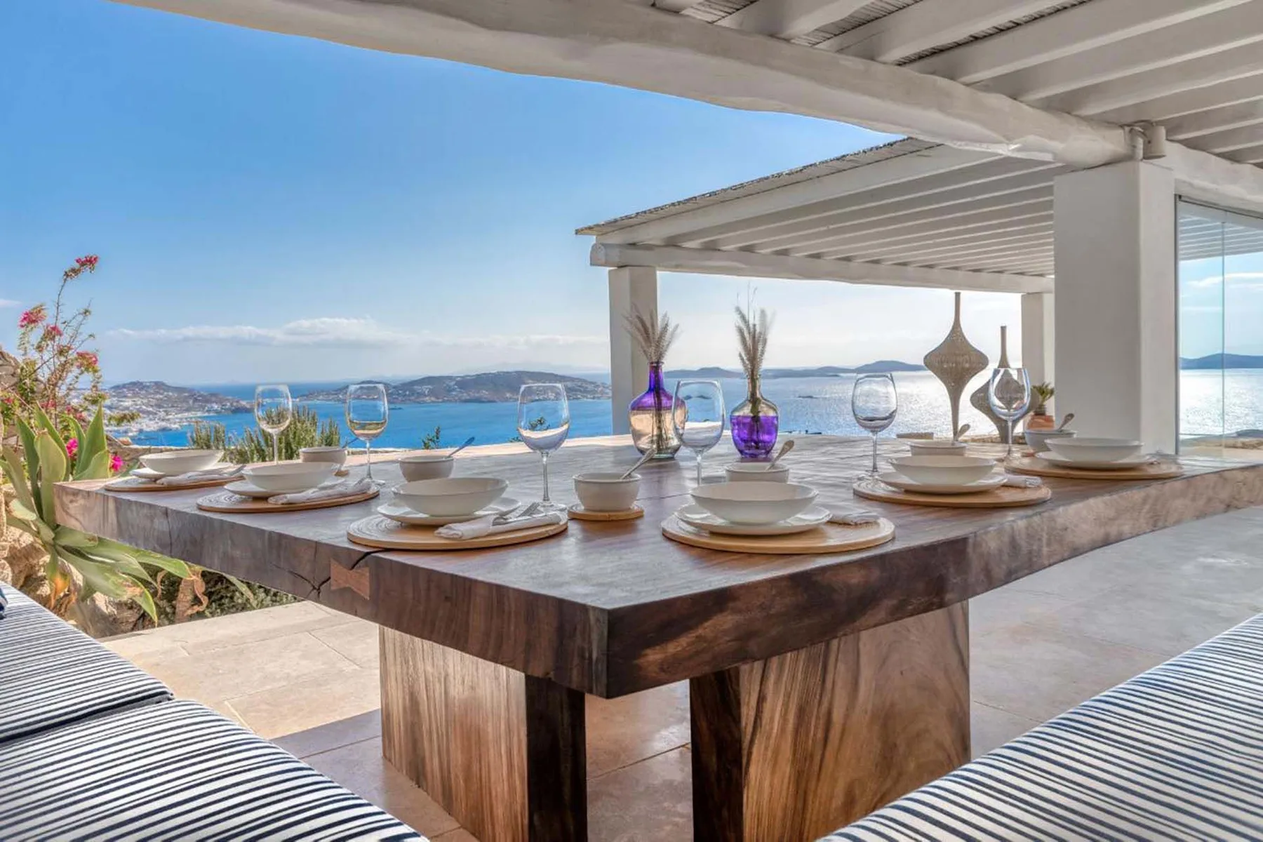 An outdoor dining table is set with glassware beside a view of the sea.
