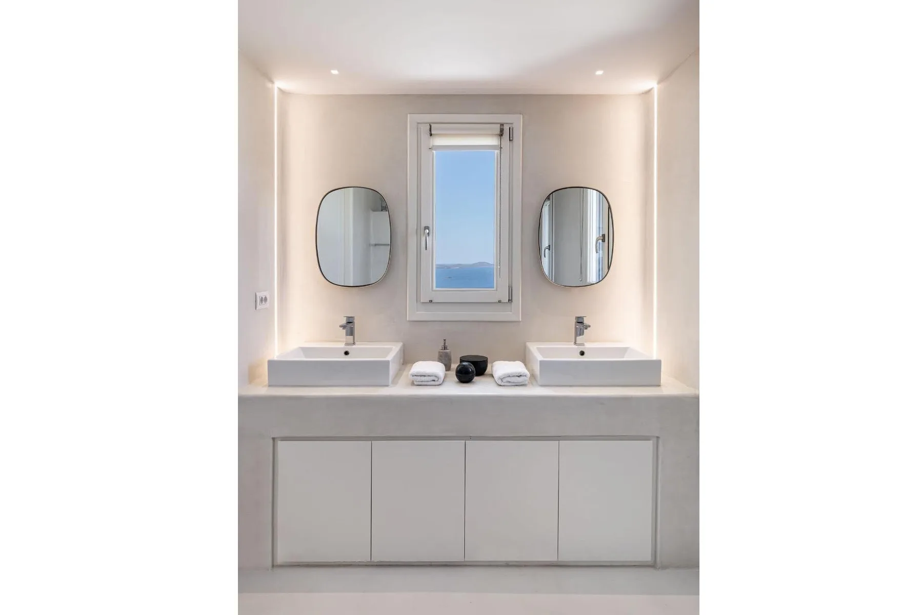 A bright bathroom vanity has two sinks, mirrors, and a window overlooking the water.