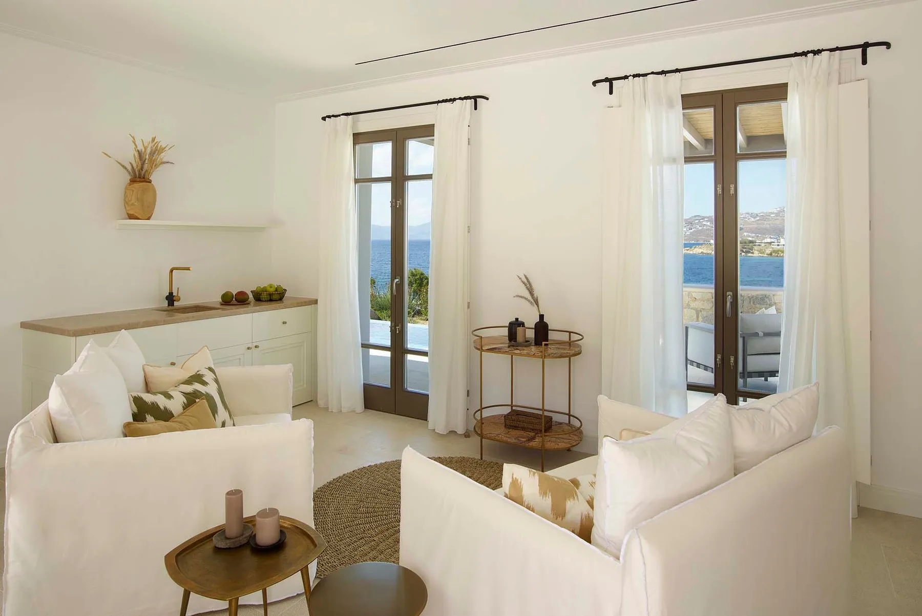 A bright living room with seating and tall windows overlooking the sea.