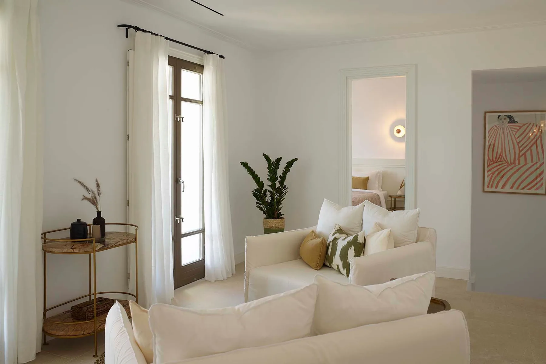 A bright living room with pale seating, a tall window, and a potted plant.