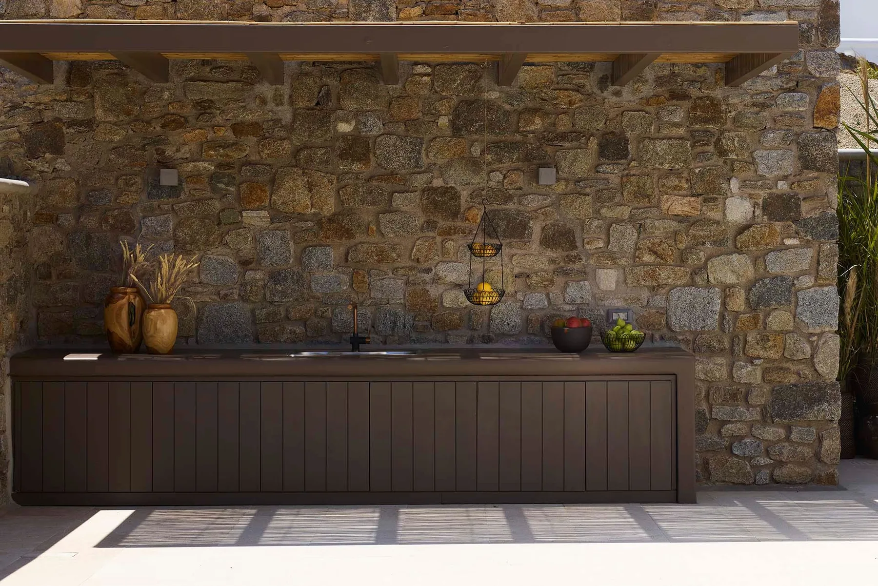 An outdoor kitchen counter stands against a stone wall beneath a canopy.