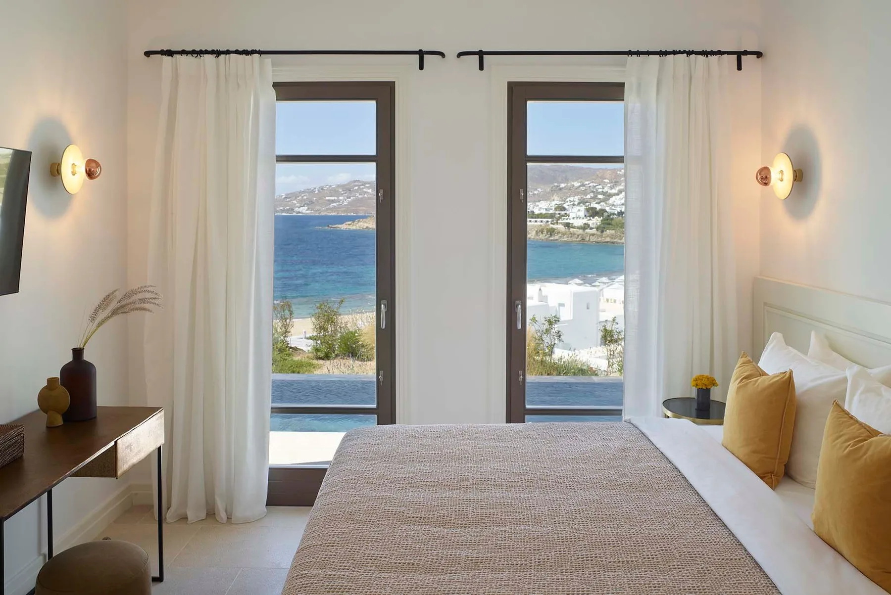 A bedroom with a bed beside two tall windows overlooking the sea.