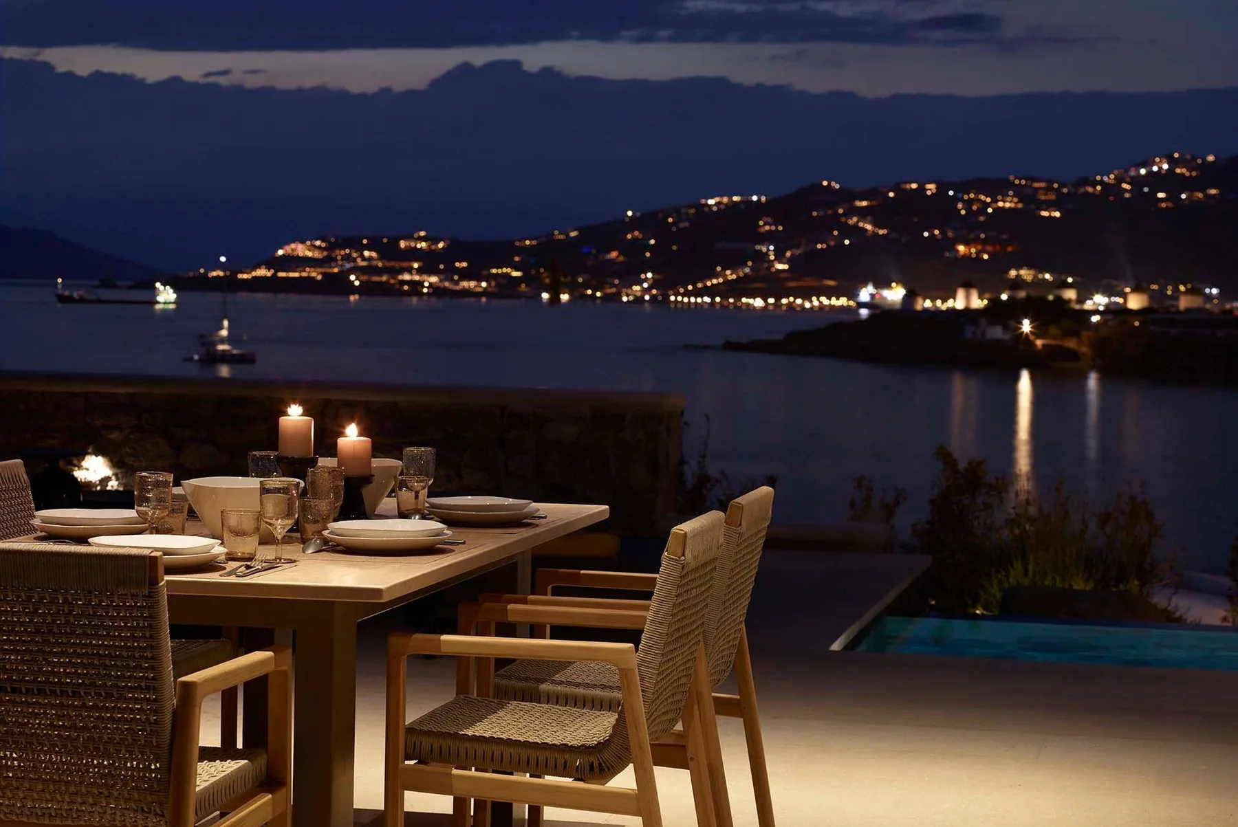 An outdoor dining area overlooks a bay and illuminated hillside at dusk.