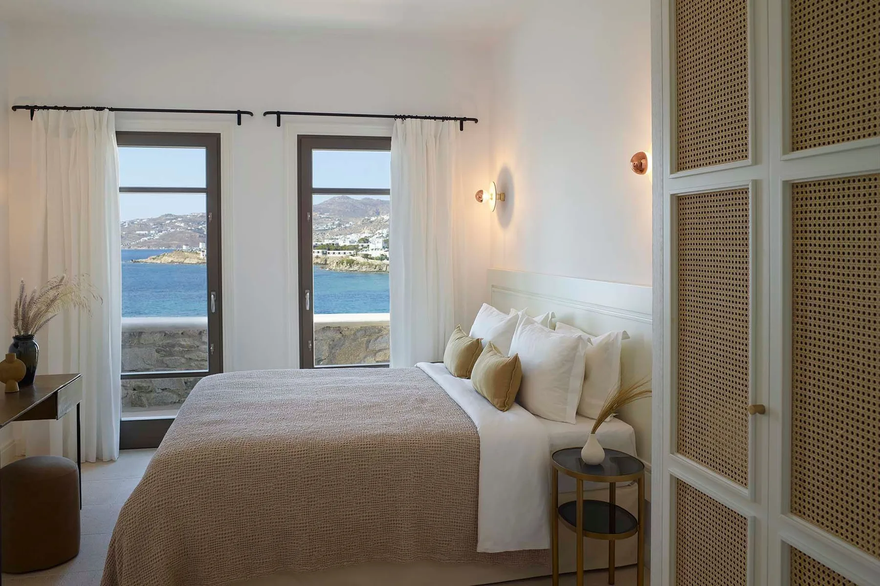 A bright bedroom with a neatly made bed and windows overlooking the water.