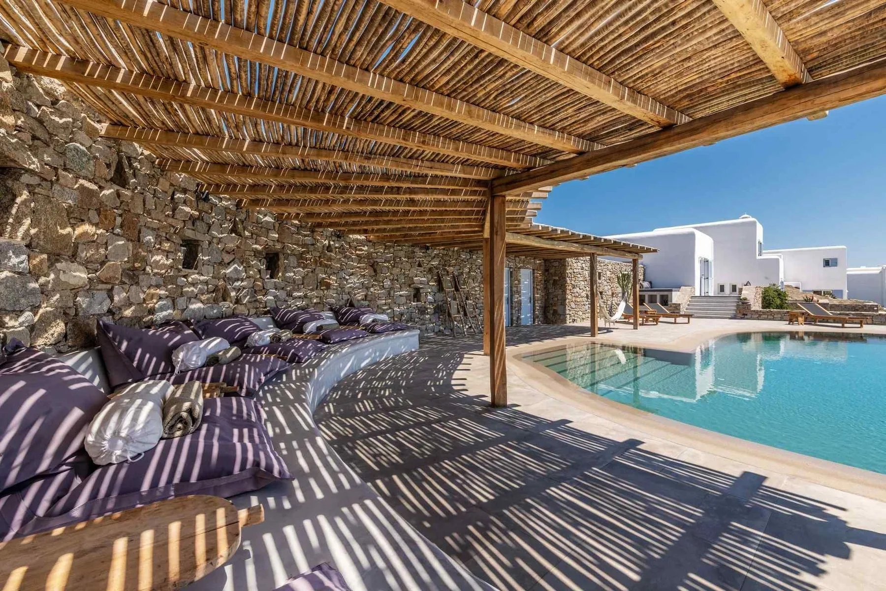 A shaded poolside lounge with cushioned seating beneath a wooden pergola.