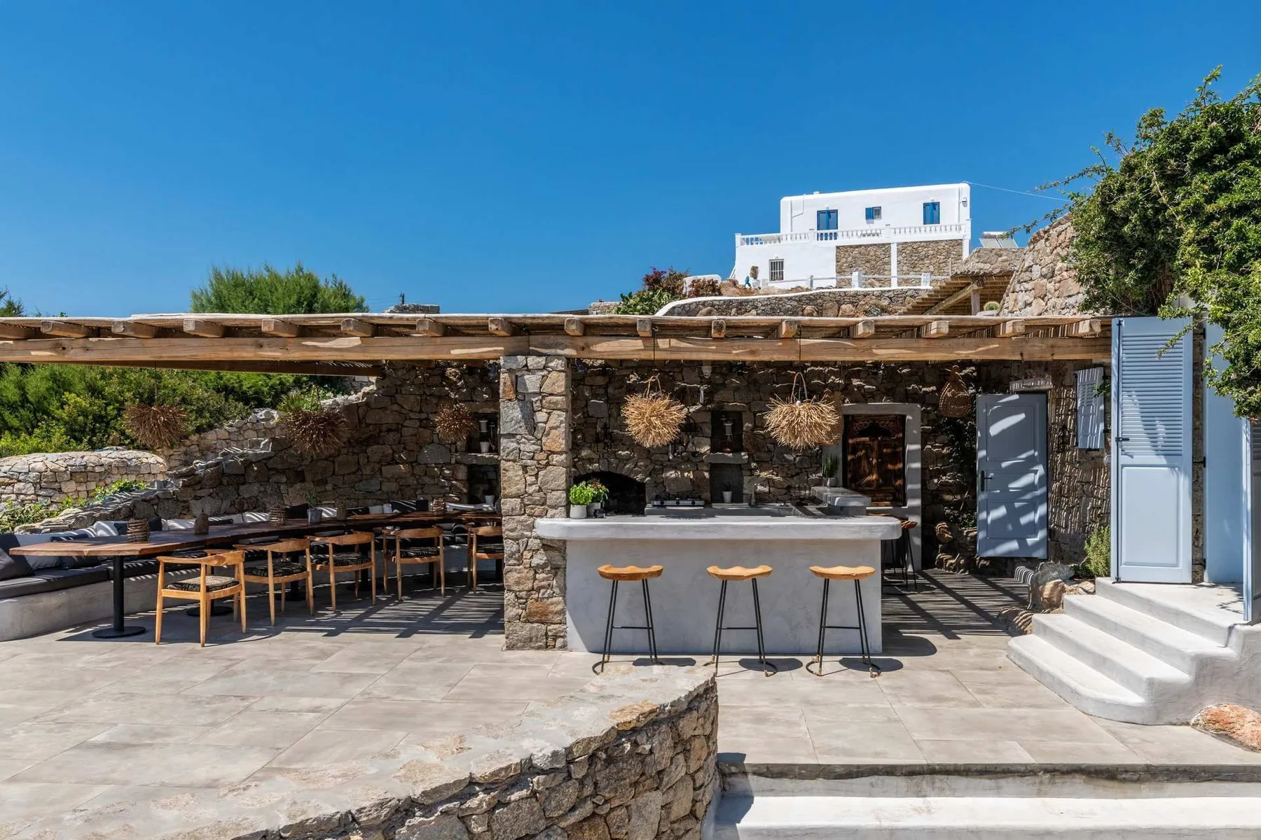 An outdoor stone patio with a bar counter and dining table beneath a wooden pergola.
