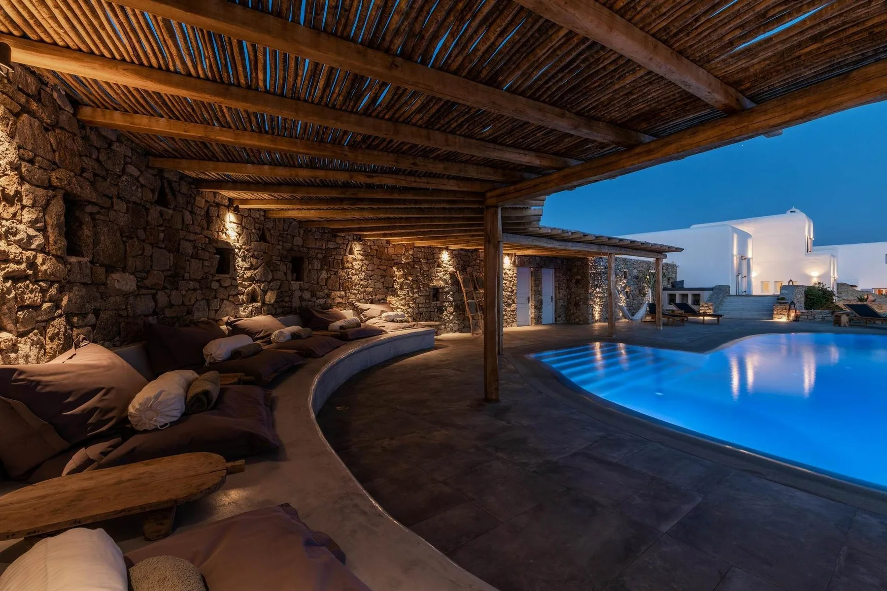 A covered outdoor lounge beside a lit swimming pool at dusk.