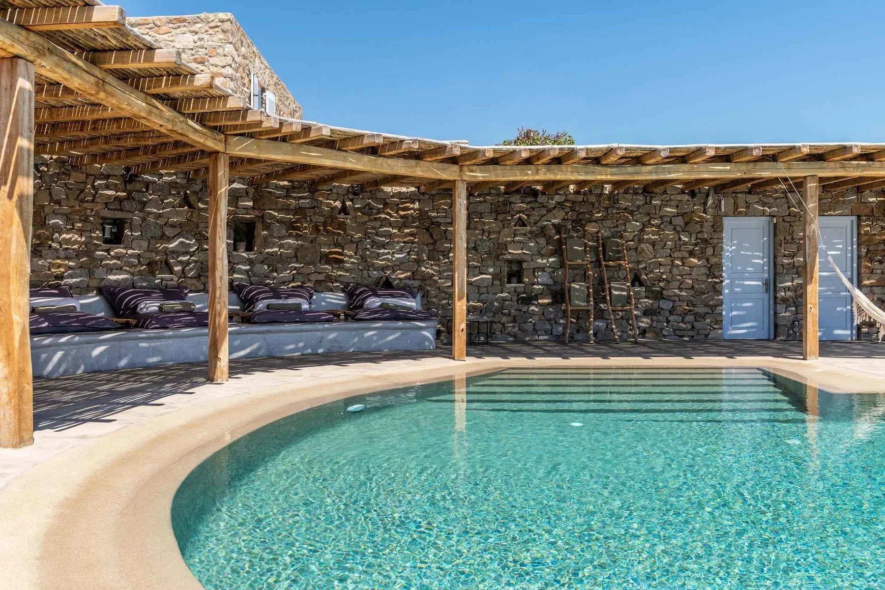 An outdoor swimming pool sits beside a shaded lounge area and stone wall.