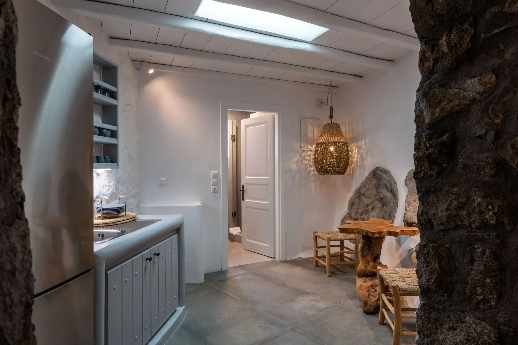 A bright kitchen opens into a dining area with a wooden table and woven pendant light.