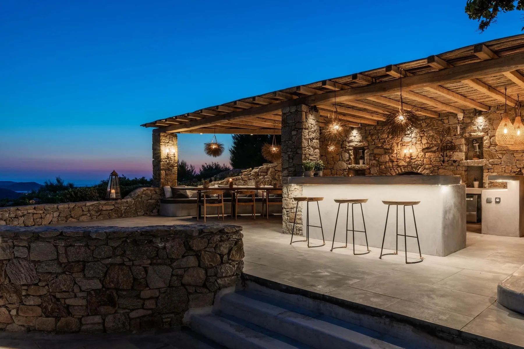 An outdoor stone bar beneath a wooden pergola, with stools and warm hanging lights.