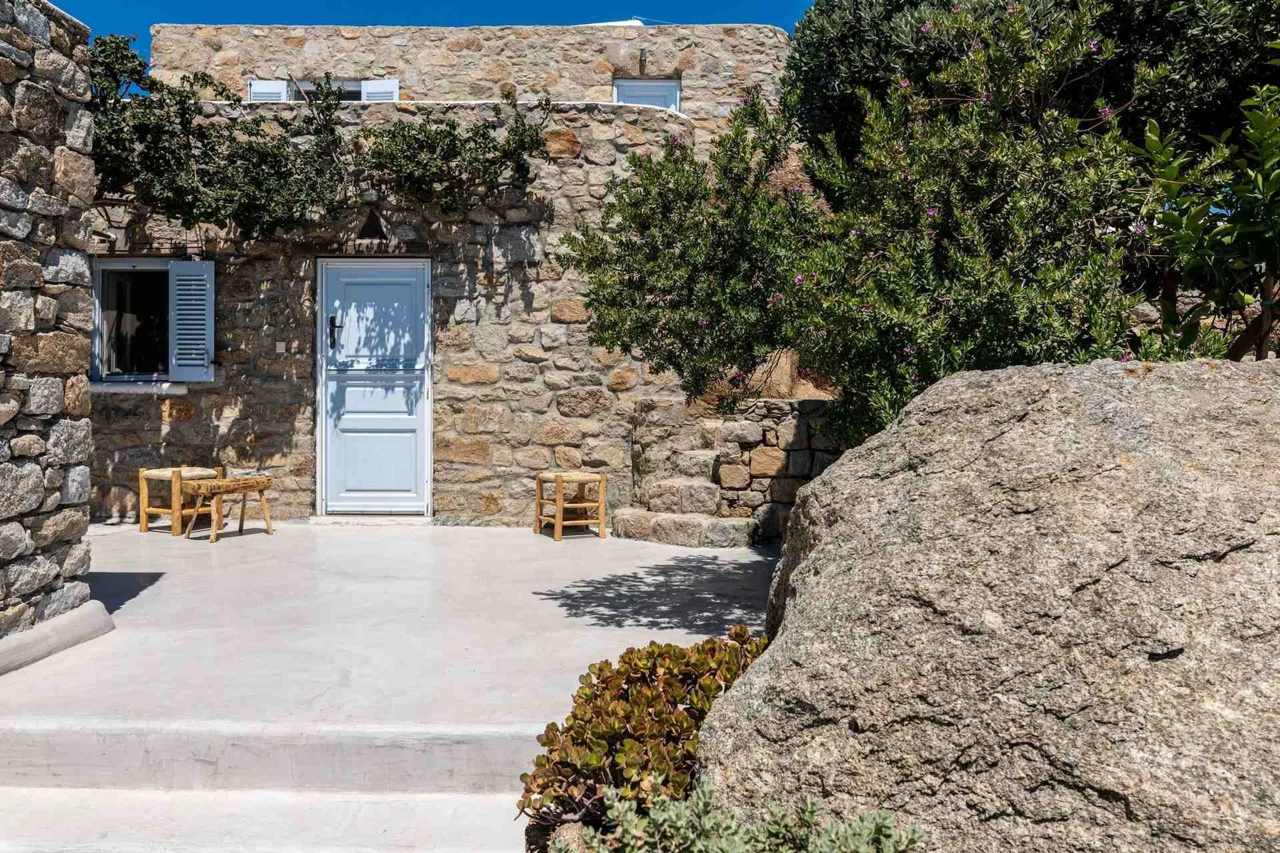A stone courtyard with a pale door, shaded seating, and a large rock.