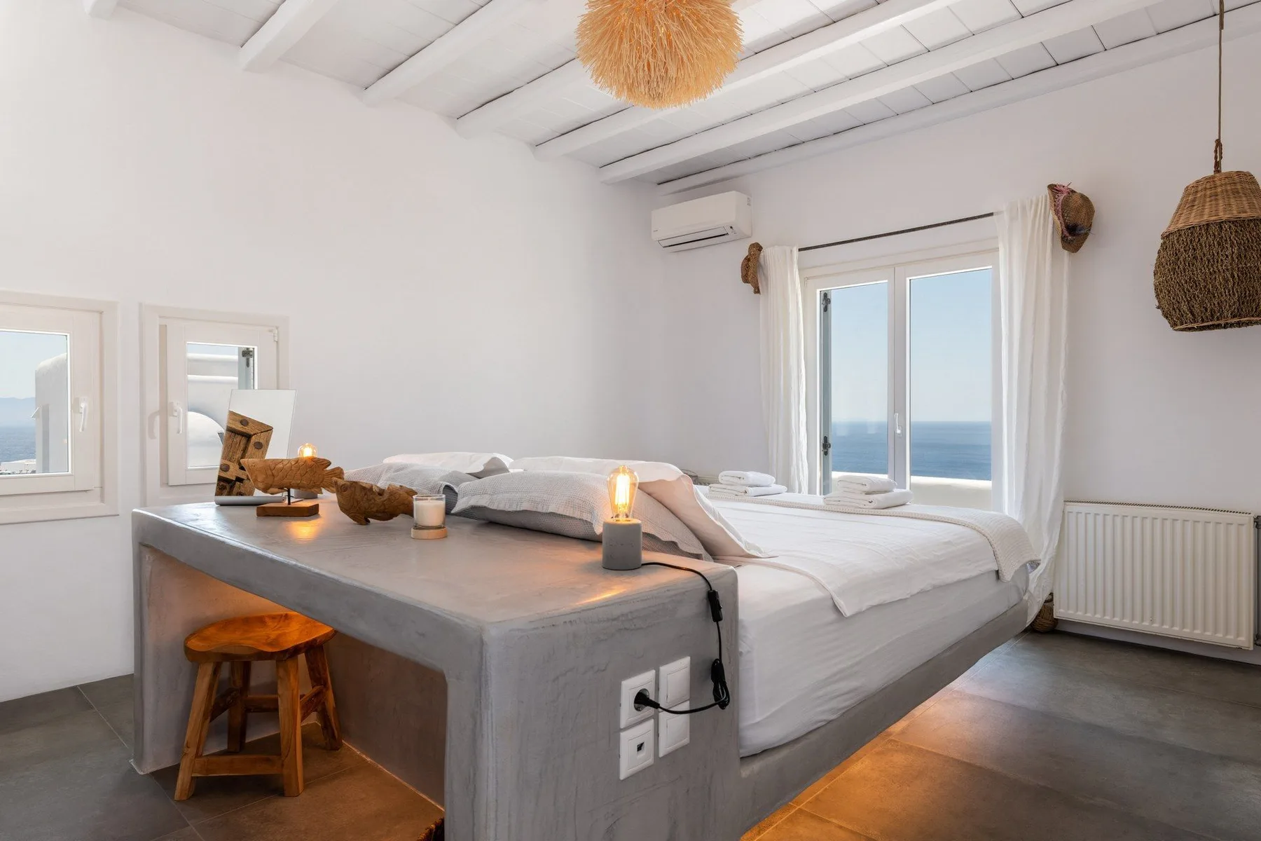A bright bedroom with a bed beside large windows overlooking the sea.