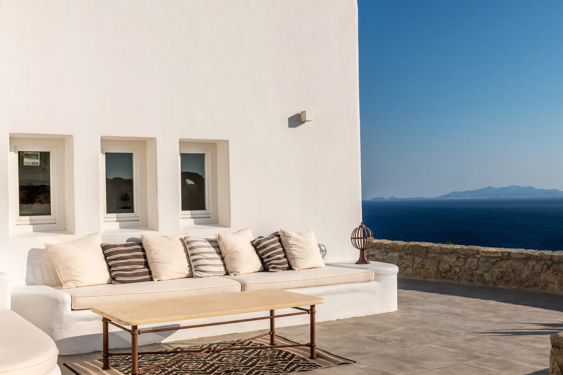 A white outdoor seating area faces the sea beneath a clear blue sky.