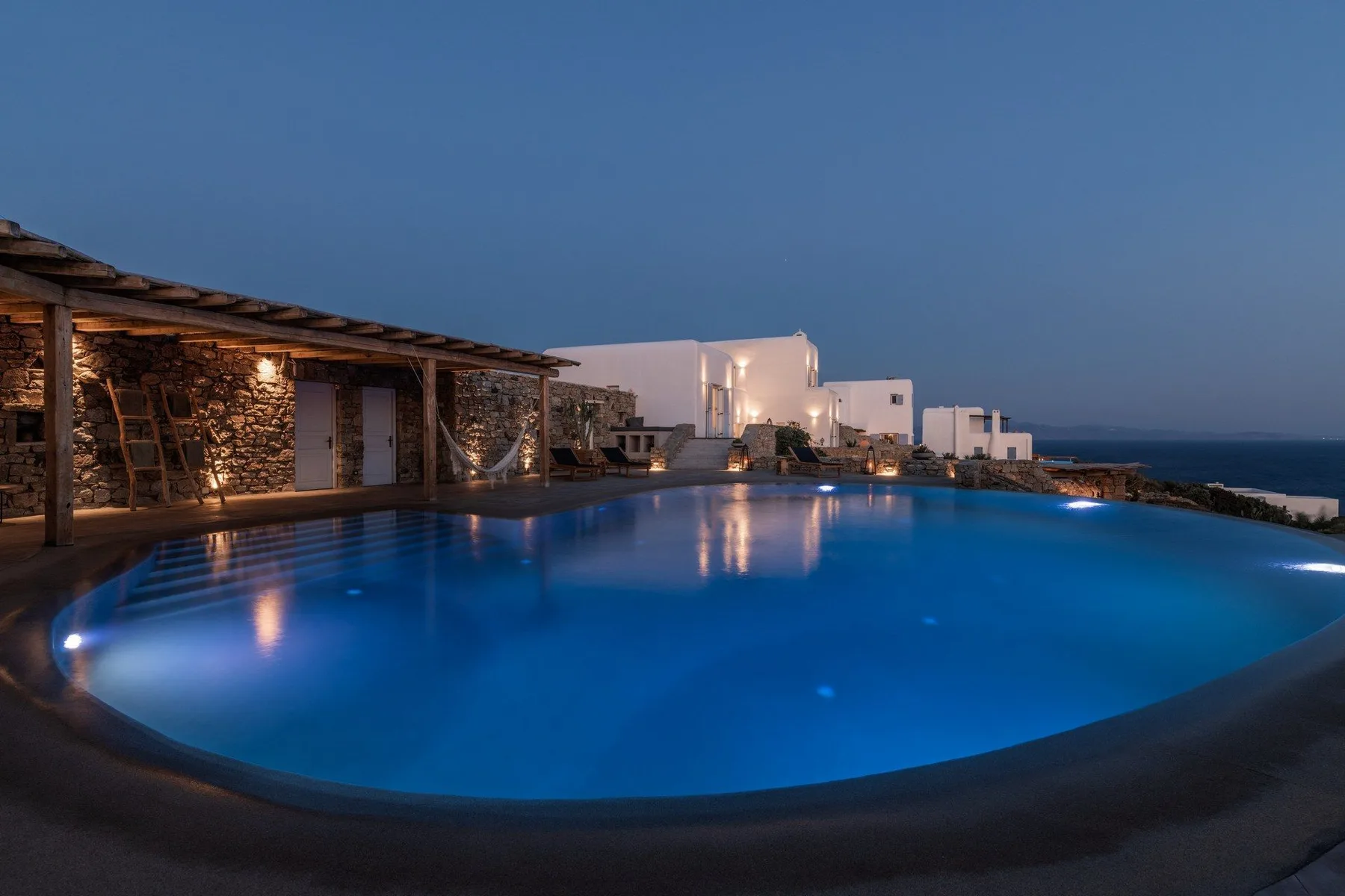 An illuminated outdoor pool sits beside stone buildings overlooking the sea at dusk.