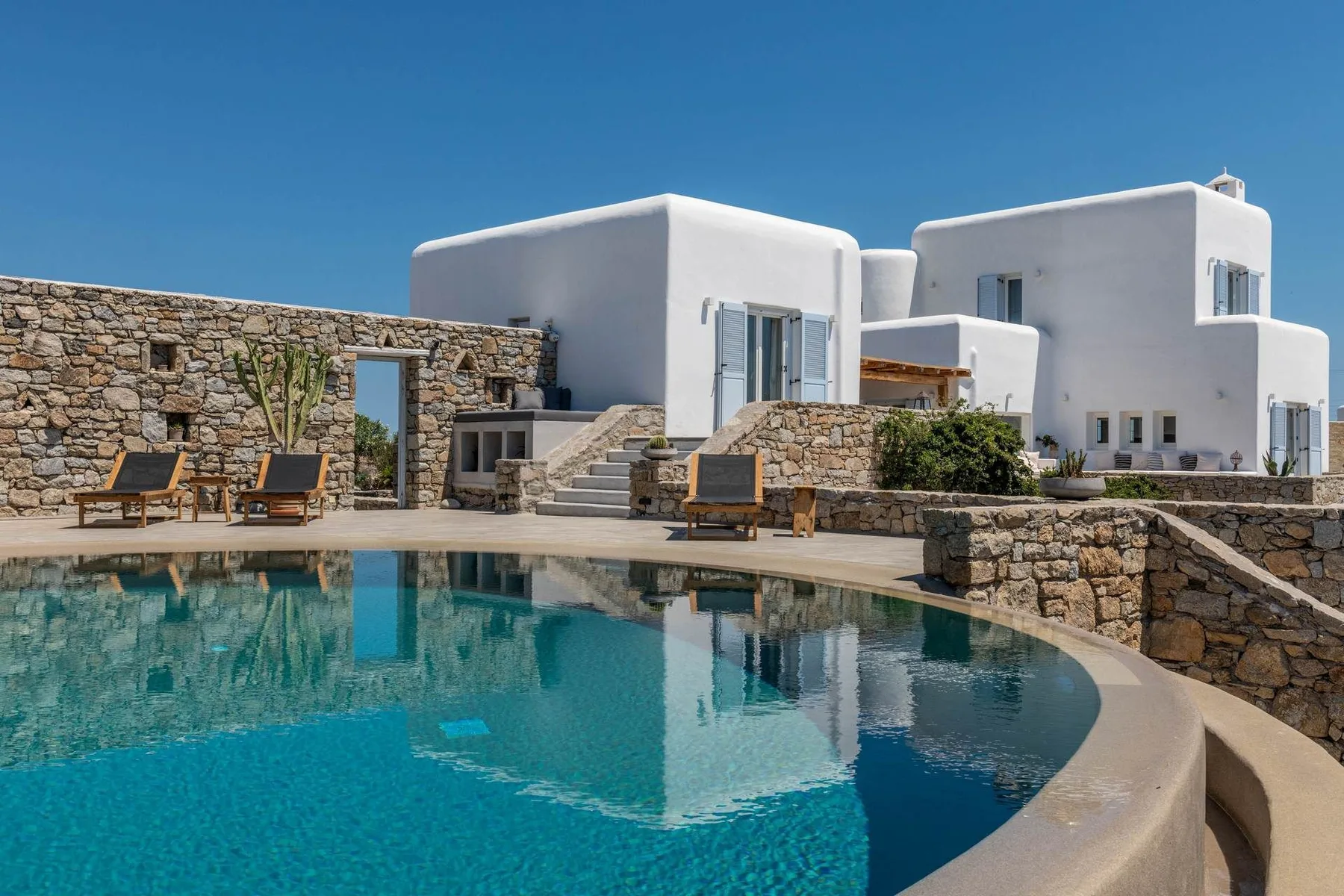 A swimming pool sits beside white buildings and stone walls under a clear blue sky.