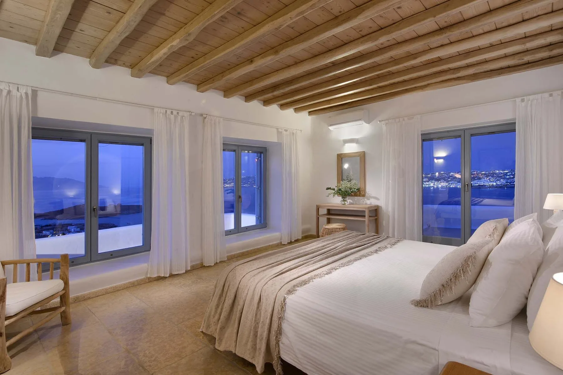 A bedroom with a bed, exposed wooden ceiling beams, and windows overlooking a lit landscape.