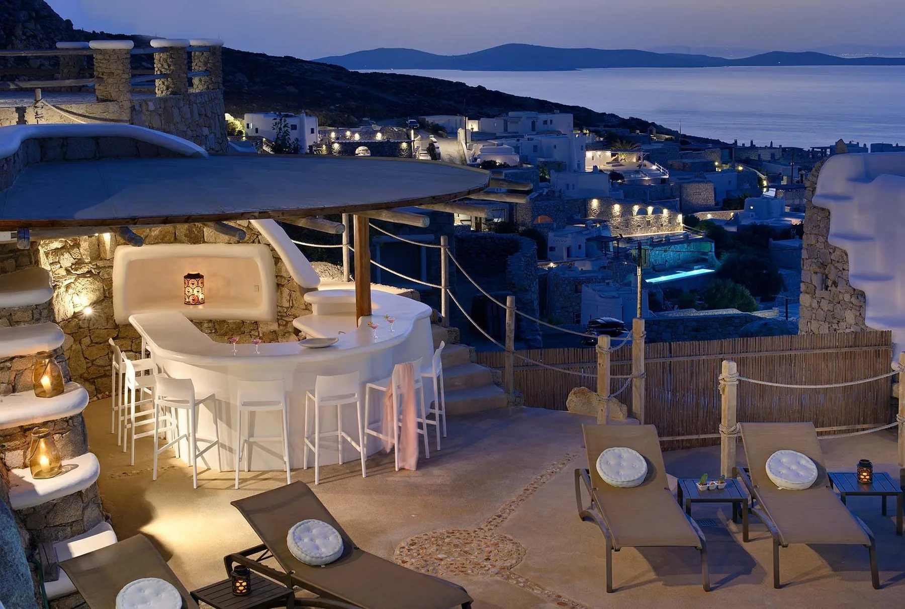 An outdoor terrace dining area overlooks a coastal town and the sea at dusk.