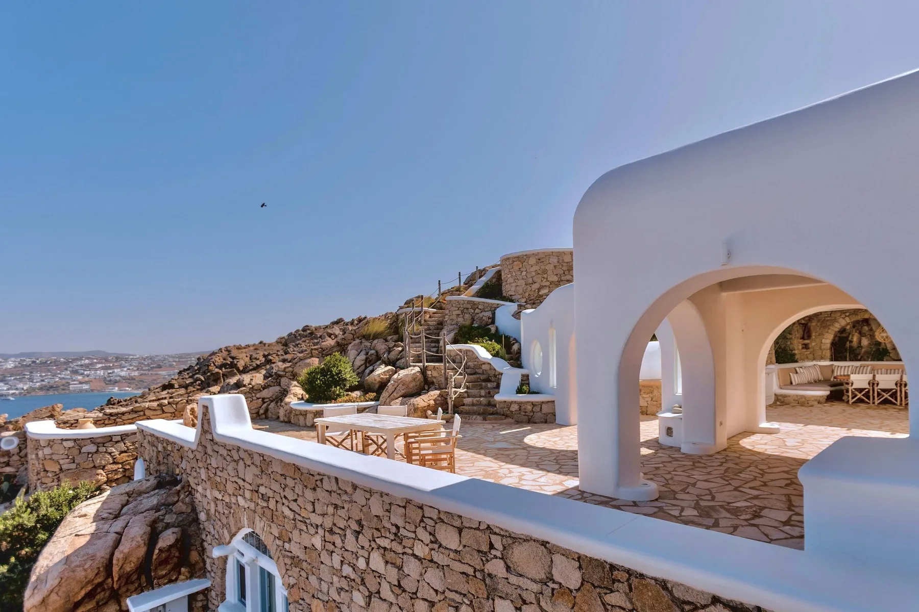 A white arched terrace overlooks a rocky hillside and the sea.