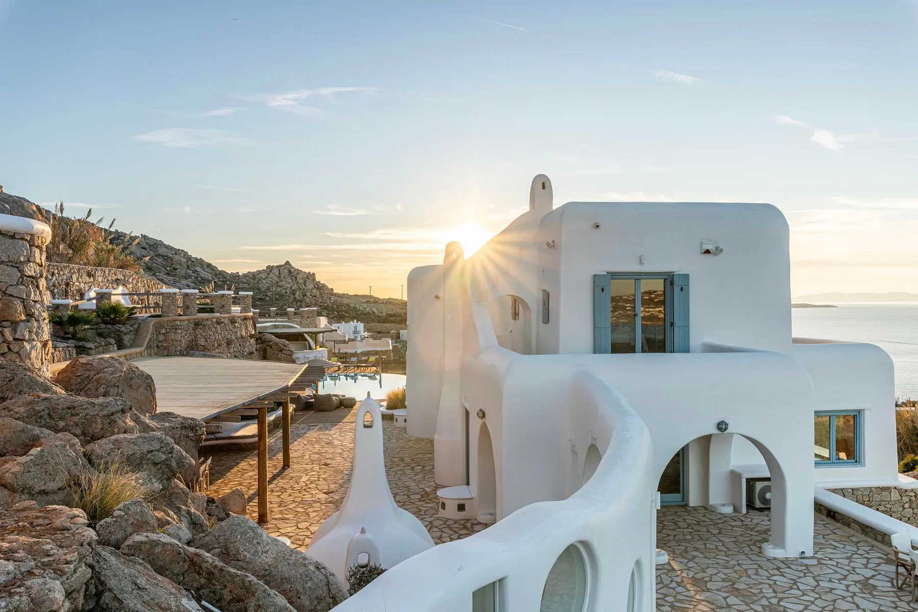 Whitewashed buildings and a stone terrace overlook a rocky coastal landscape at sunset.