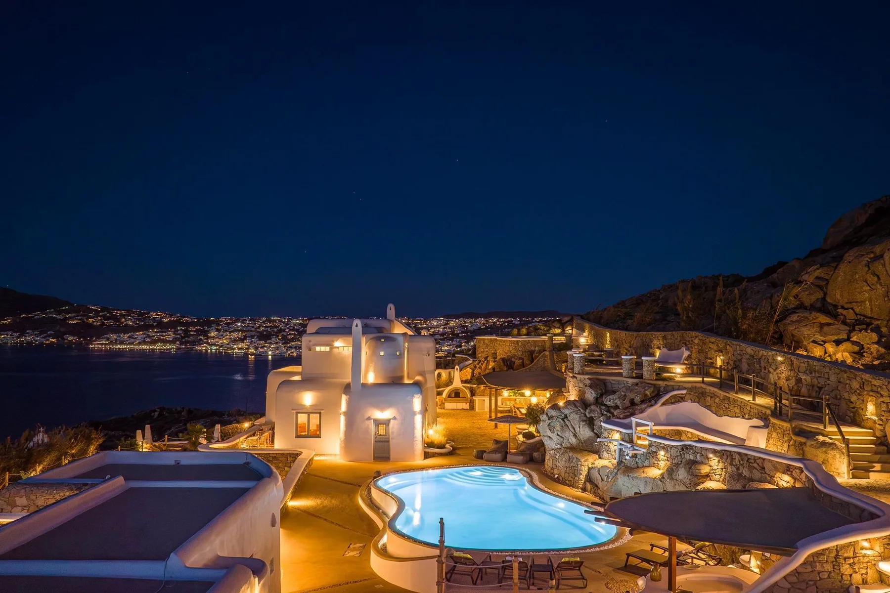 A hillside resort pool glows at night beside illuminated buildings and rocky terrain.