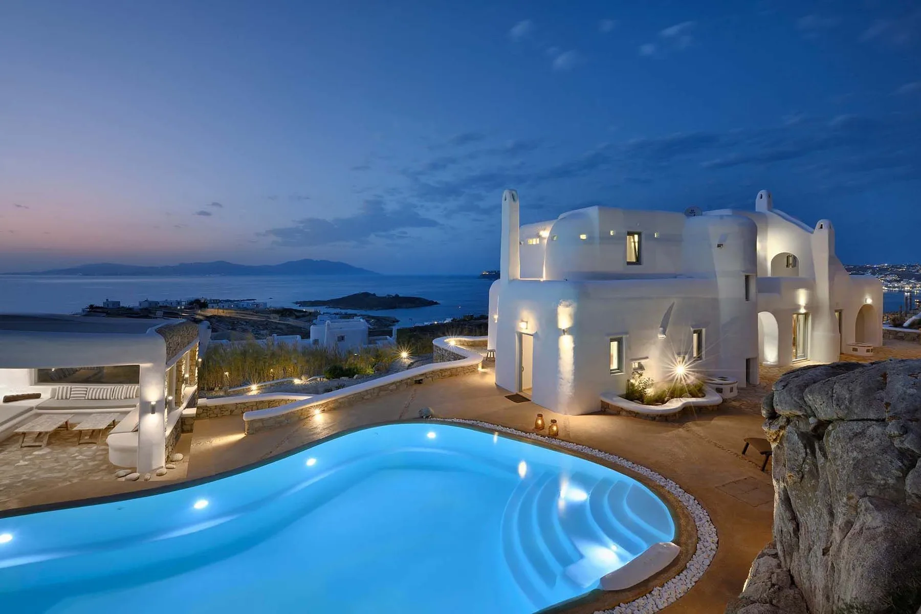 A lit white villa overlooks a curved swimming pool and the sea at dusk.