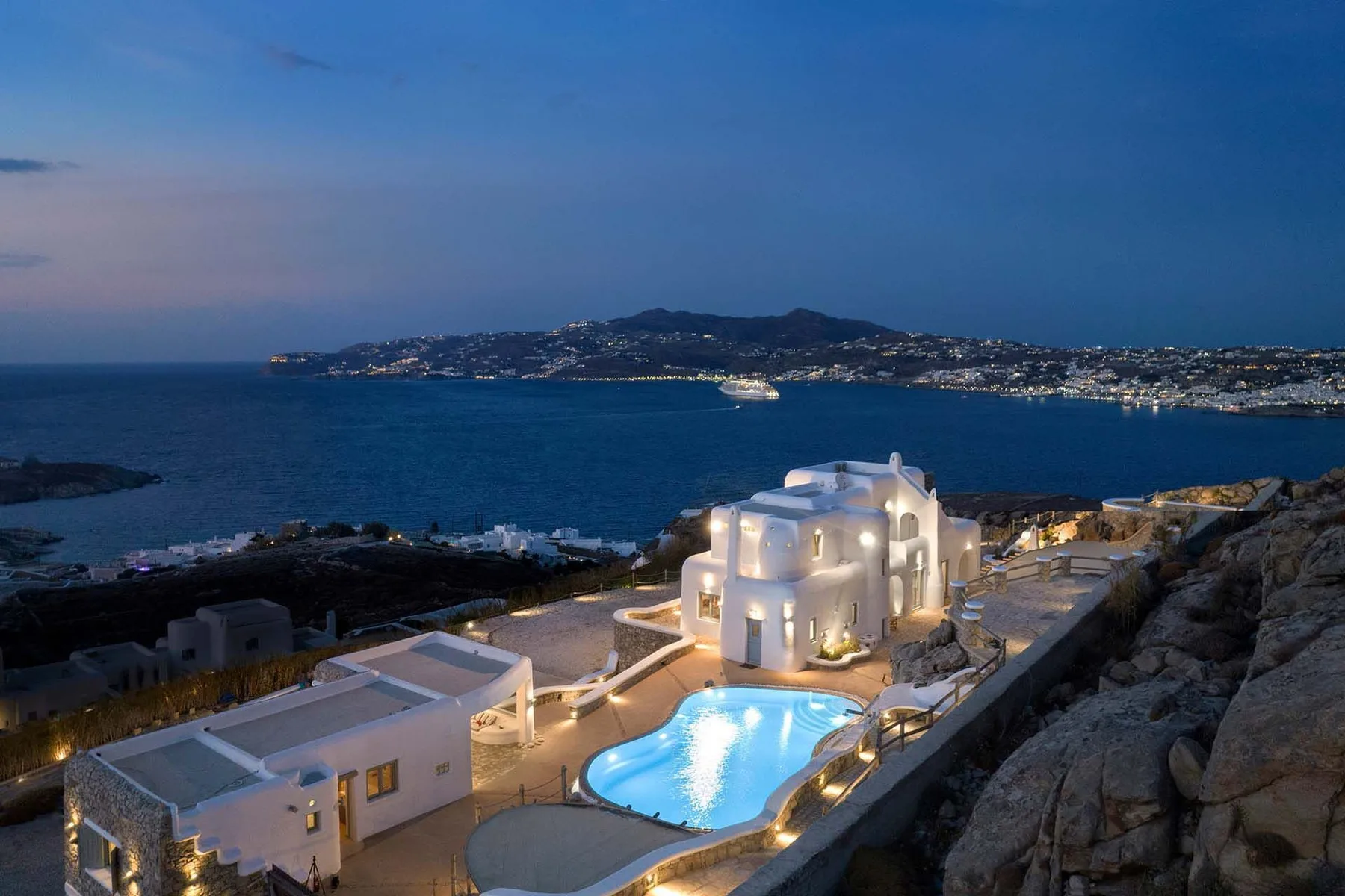A lit seaside villa overlooks a swimming pool and the bay at dusk.