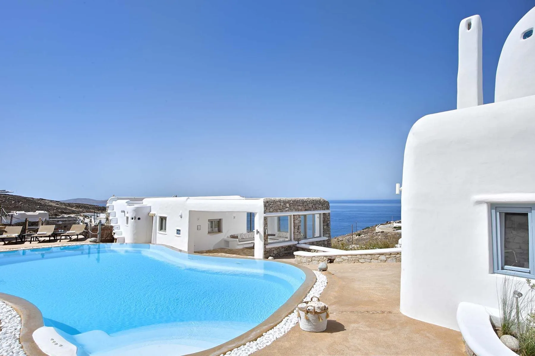 A bright outdoor swimming pool beside white buildings, with the sea visible in the distance.