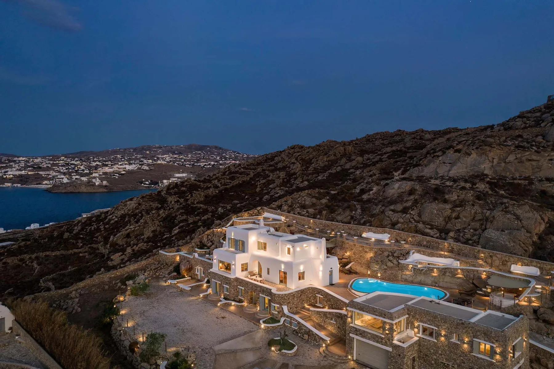 A brightly lit resort complex sits on a rocky hillside above a bay at dusk.