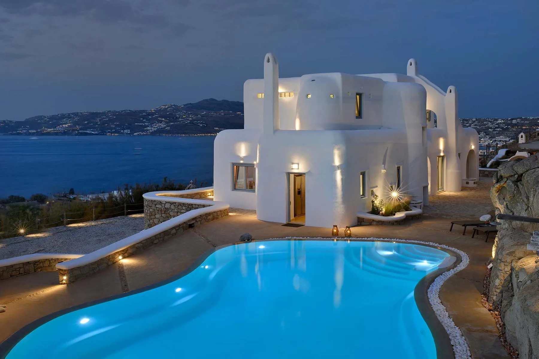 A white seaside building overlooks a brightly lit swimming pool at dusk.