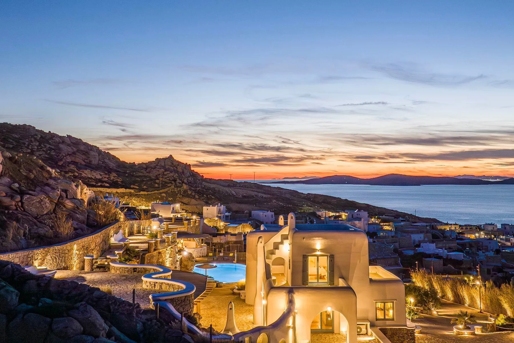 A hillside resort glows at sunset, with illuminated buildings and a pool.
