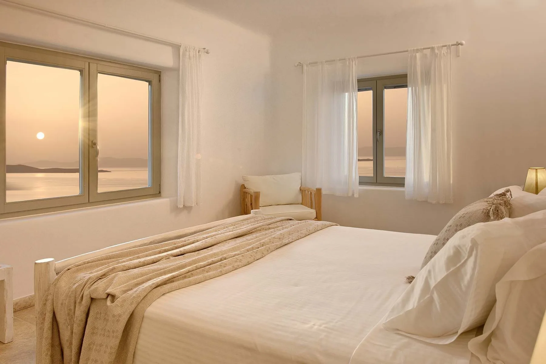 A bright bedroom with a neatly made bed and windows overlooking the water.