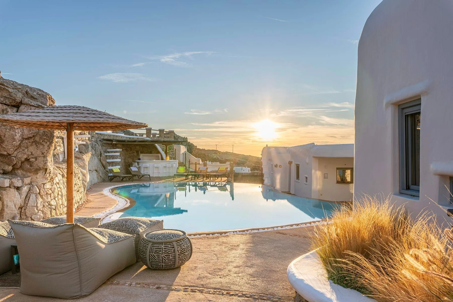 A swimming pool beside white buildings, with the sun setting in the background.
