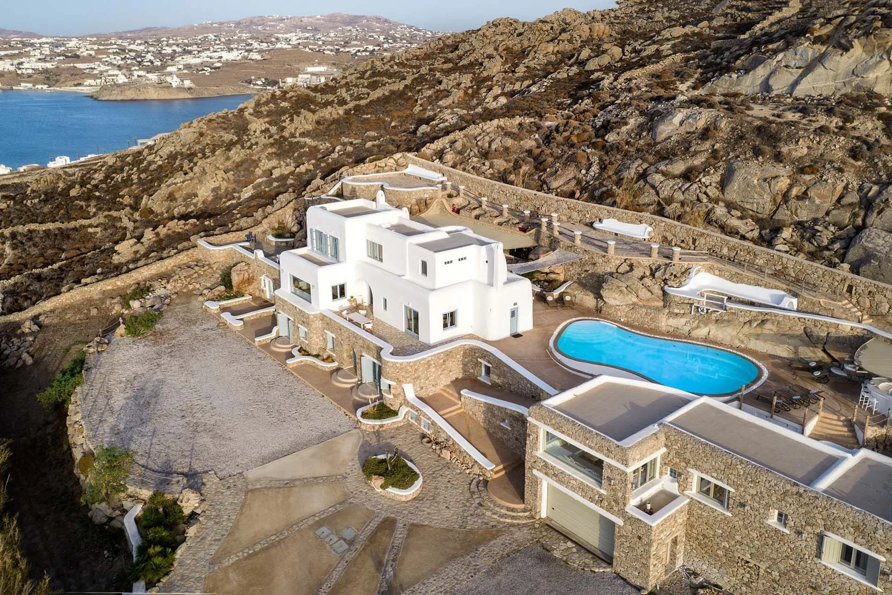 A white villa with a blue swimming pool sits on a rocky hillside overlooking the sea.