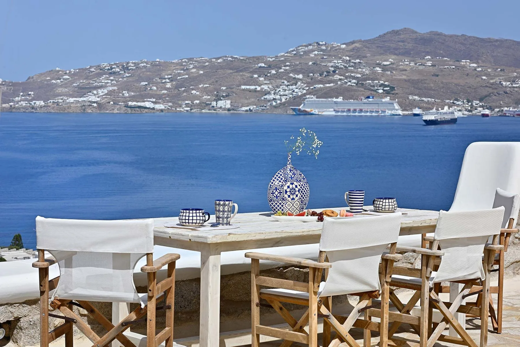 An outdoor dining area overlooks the blue sea and a hillside dotted with buildings.