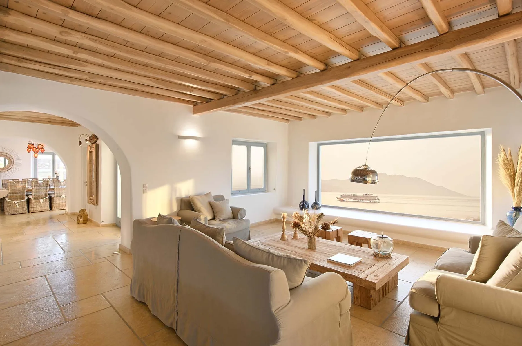 A bright living room has a seating area beside a large window overlooking the water.