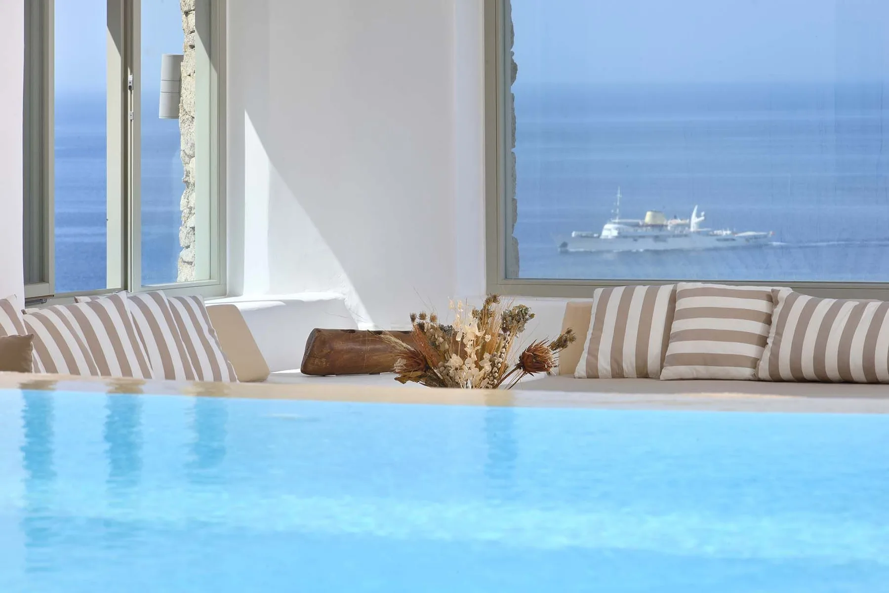 A bright indoor pool sits beside striped seating and large windows overlooking the sea.