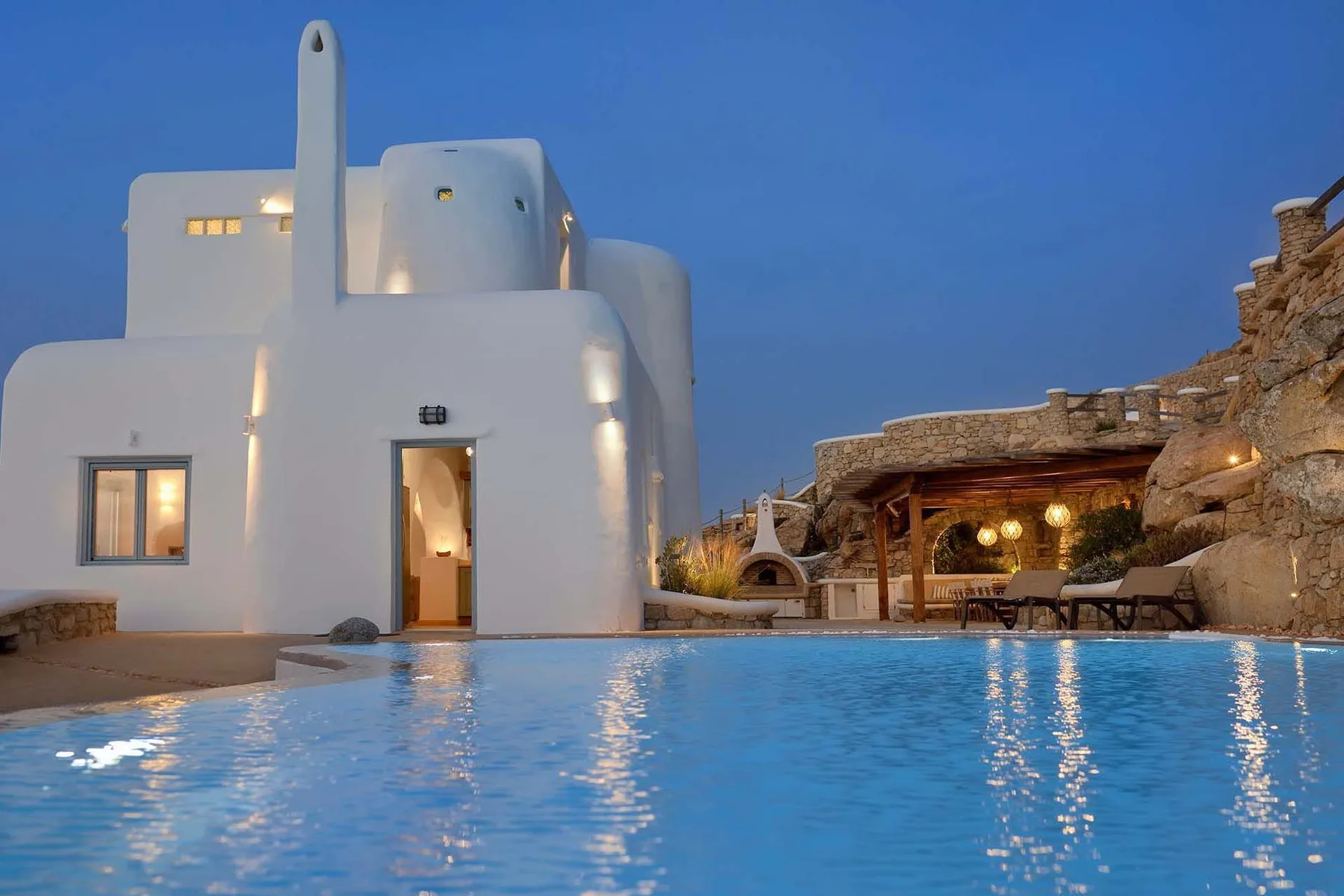 A swimming pool in front of a white building and a stone-covered terrace at dusk.
