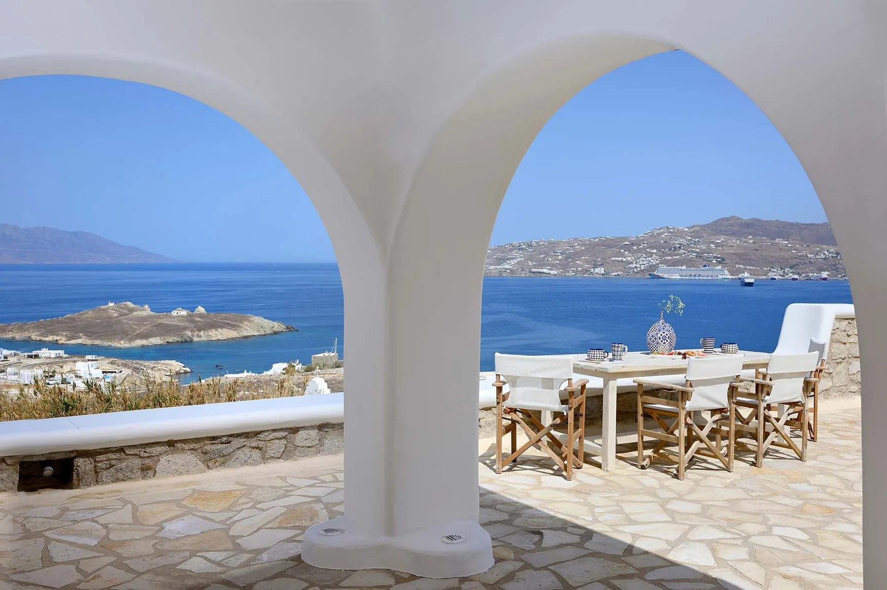 A seaside terrace with dining seating overlooks blue water through two arches.