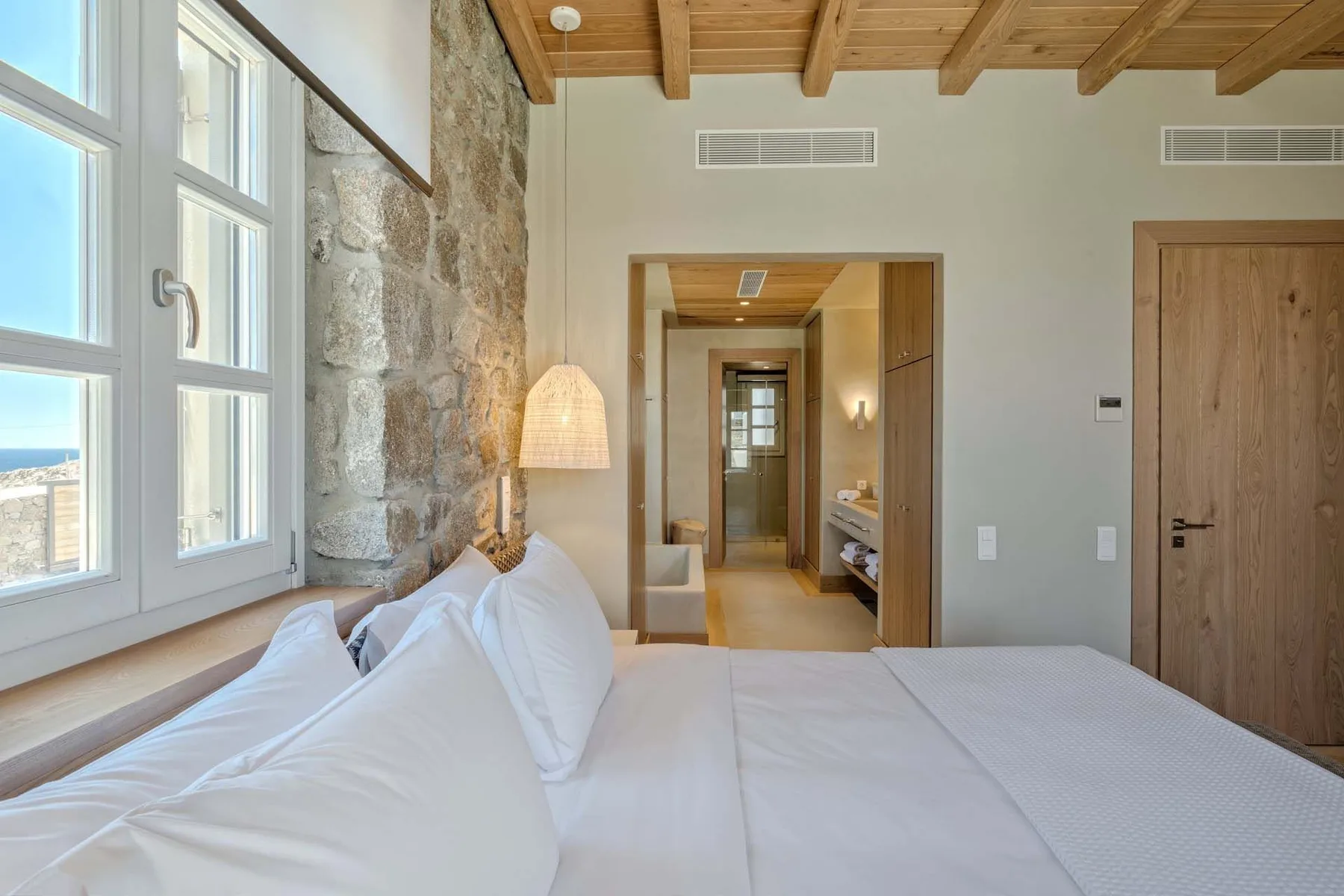 A bright bedroom with white bedding, tall windows, and an exposed stone wall.