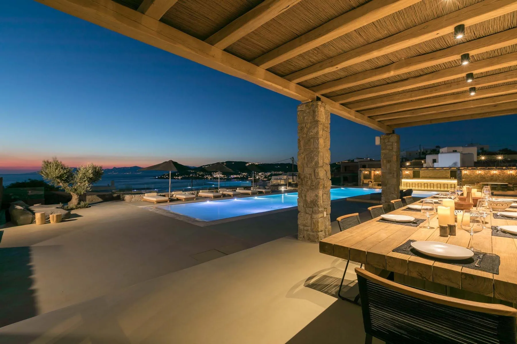 A covered outdoor dining area overlooks a lit swimming pool and distant hills at dusk.