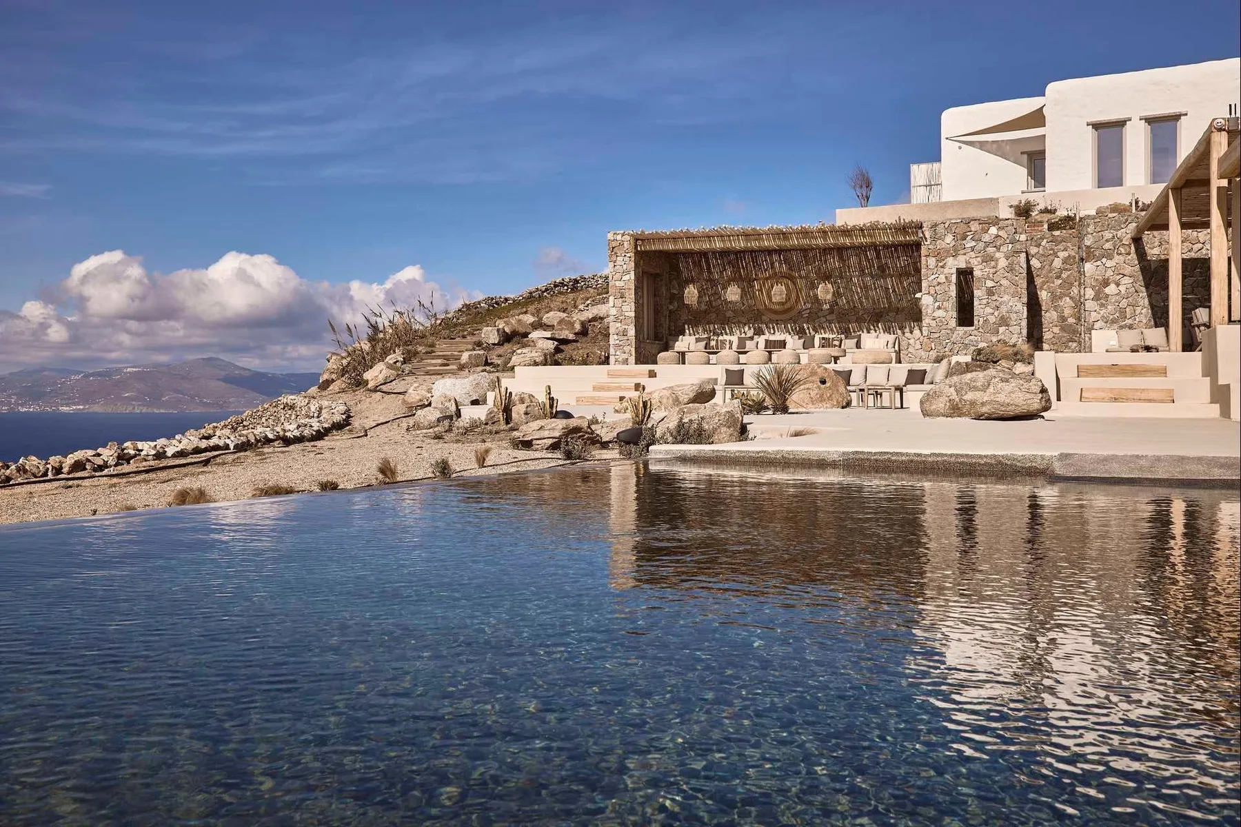 A seaside pool fronts a stone terrace and white buildings.