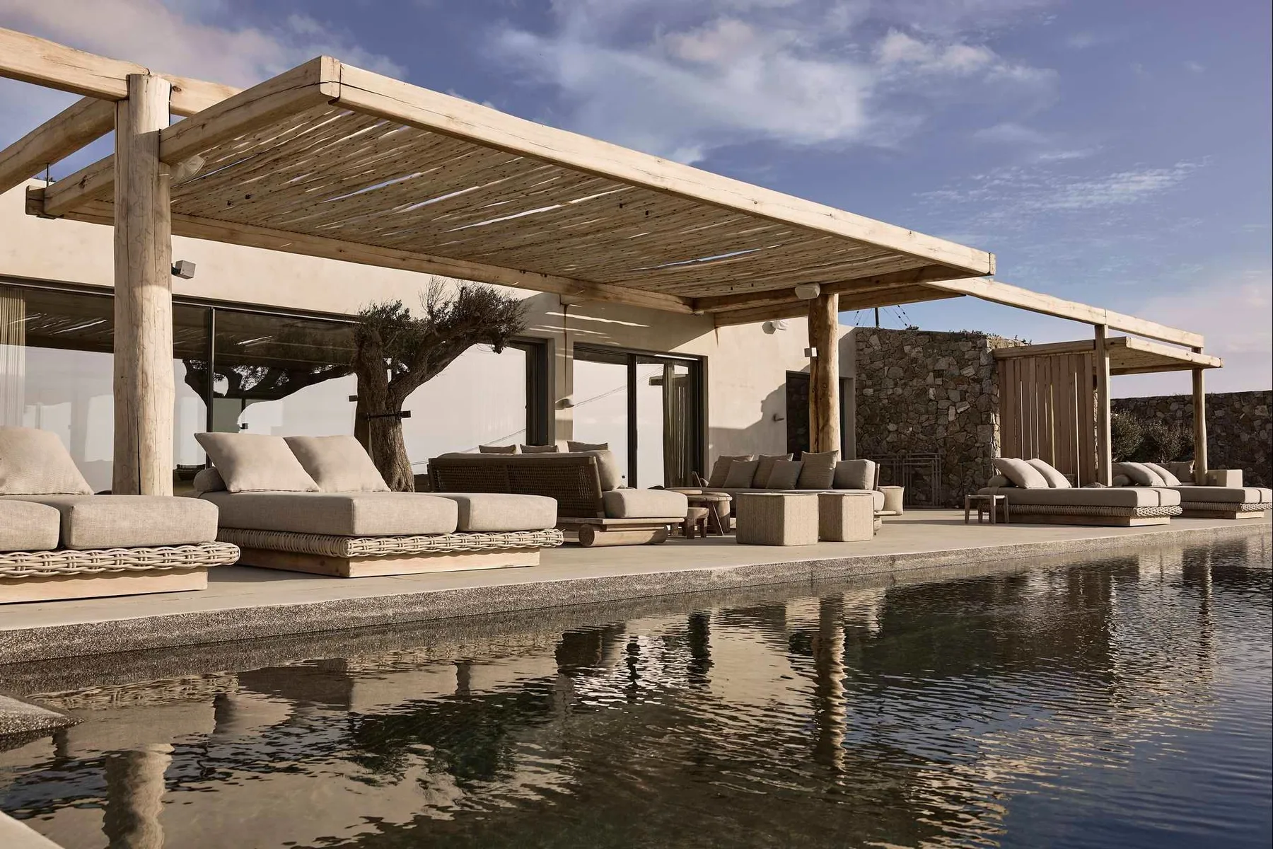 An outdoor lounge beside a swimming pool, with cushioned seating beneath a wooden pergola.