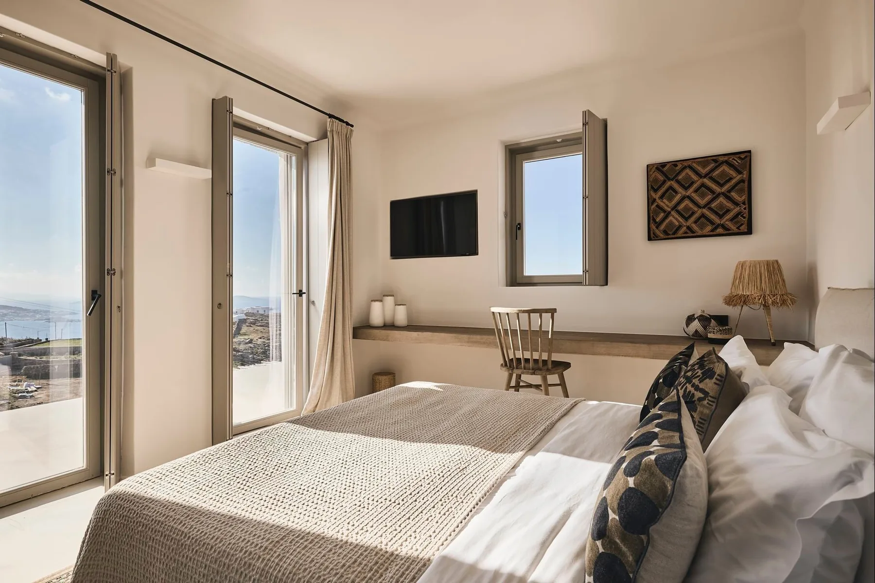 A bright bedroom with a bed, a wall-mounted television, and large windows.